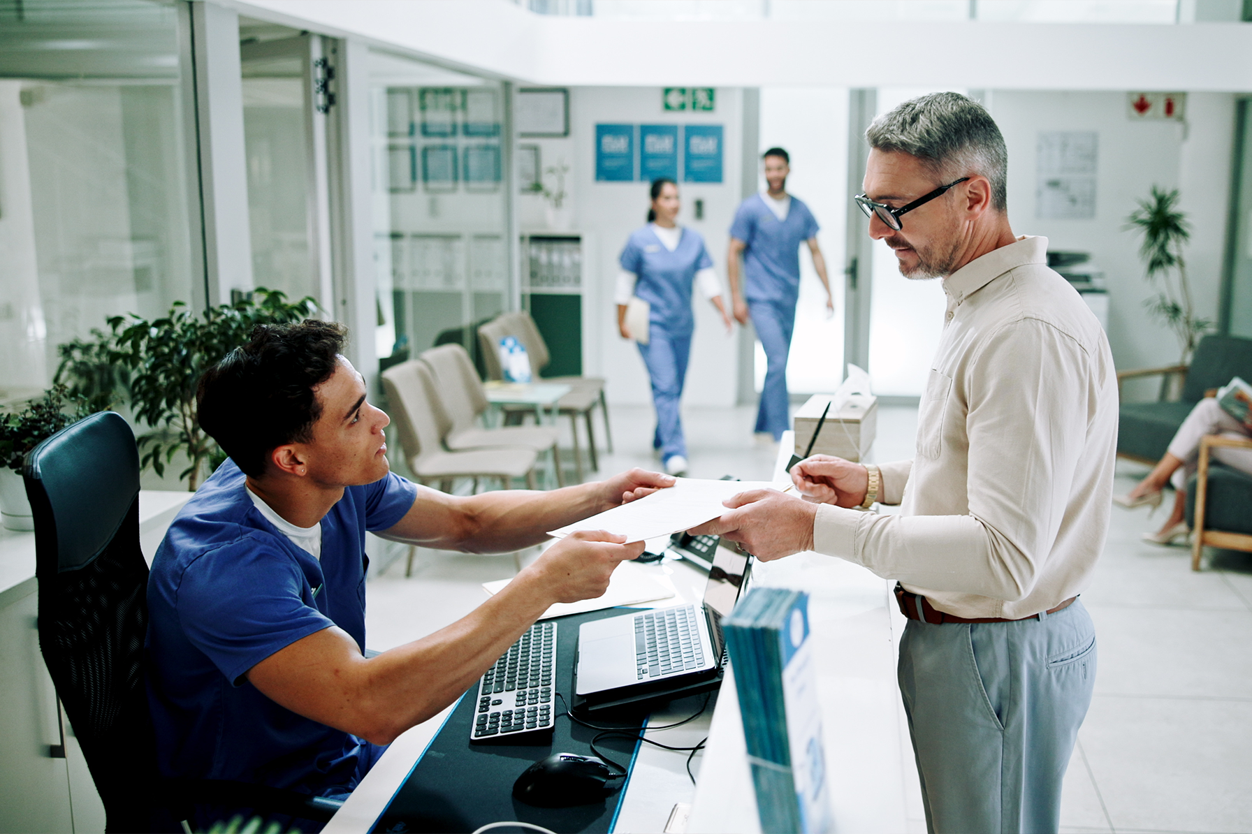 Front desk medical professional hands paperwork to a male patient in a healthcare clinic reception area. In the background, two nurses walk through the hallway, and another patient is seated in the waiting area. The setting is clean and modern, emphasizing organized patient engagement.