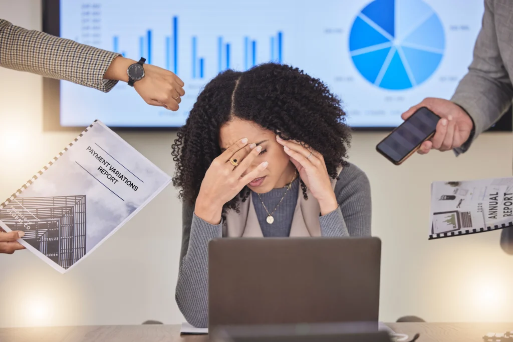 A healthcare professional sits overwhelmed at a desk with her head in her hands, surrounded by colleagues thrusting documents — including a Payment Variations Report and an Annual Report — a phone, and a watch, with financial charts displayed on a screen behind her, illustrating the stress of managing insurance underpayments and revenue discrepancies.